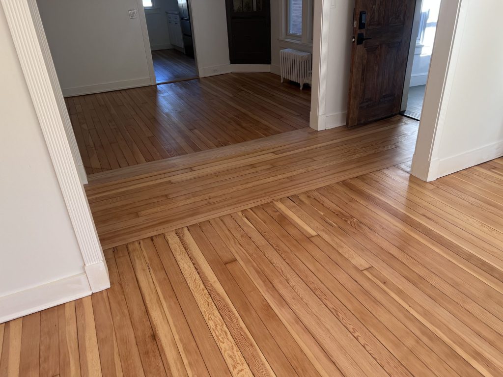 Refinished narrow-strip wood floor running through a doorway, with a band of boards laid crosswise at the transition between two rooms